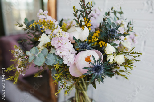 High angle of beautiful spring bouquet with assorted colorful blooming flowers including pink peonies and blue sea holly flowers with mimosa twigs arranged on glass vase on table near white wall photo