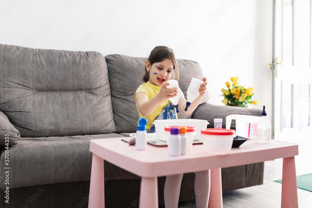 Adorable girl in casual wear with dirty face pouring liquids into plastic container from bottles while sitting on sofa and enjoying handicraft Adorable girl in casual wear with dirty face pouring liquids into plastic container from bottles while sitting on sofa and enjoying handicraft