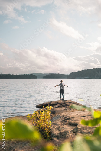 Back view of anonymous female hiker standing on stony shore near calm lake and enjoying freedom during cloudy summer day in Algonquin Provincial Park in Canada