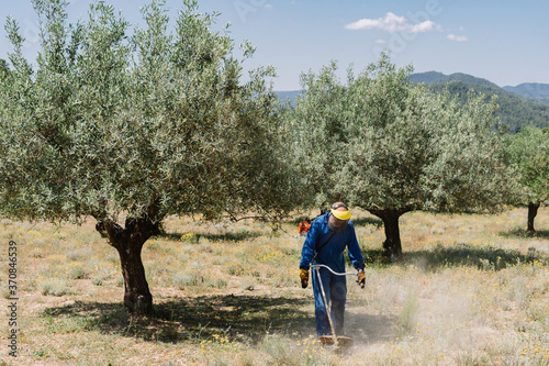 Male worker in protective helmet and uniform mowing grass with brush cutter on sunny day on background of amazing landscape