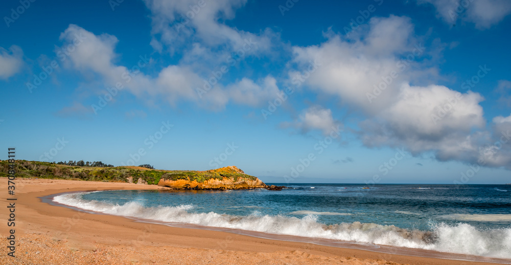 A curved orange sand beach with white waves and dramatic clouds in the blue sky