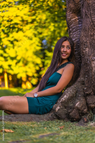 A young pretty brunette Latina with long straight hair leaning against a tree in a green dress. Sitting enjoying on the grass next to a tree in the park