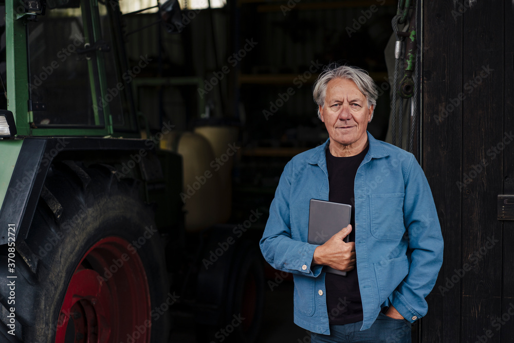 Portrait of a confident senior man holding tablet on a farm with tractor in barn Portrait of a confident senior man holding tablet on a farm with tractor in barn