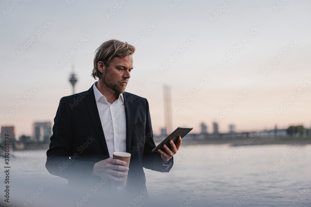 Businessman with takeaway coffee and tablet at the riverside at dusk Businessman with takeaway coffee and tablet at the riverside at dusk