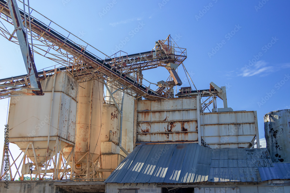 Fototapeta premium Horizontal view of conveyors, silos, hopper, and tin roofing of an old concrete plant against the background of a blue sky..