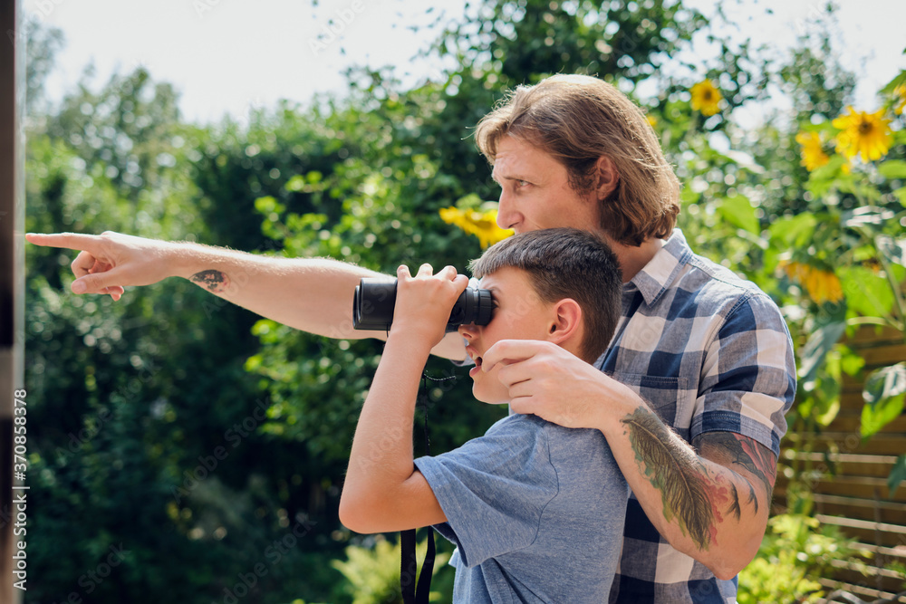 Father showing something to son looking through binoculars at yard during sunny day