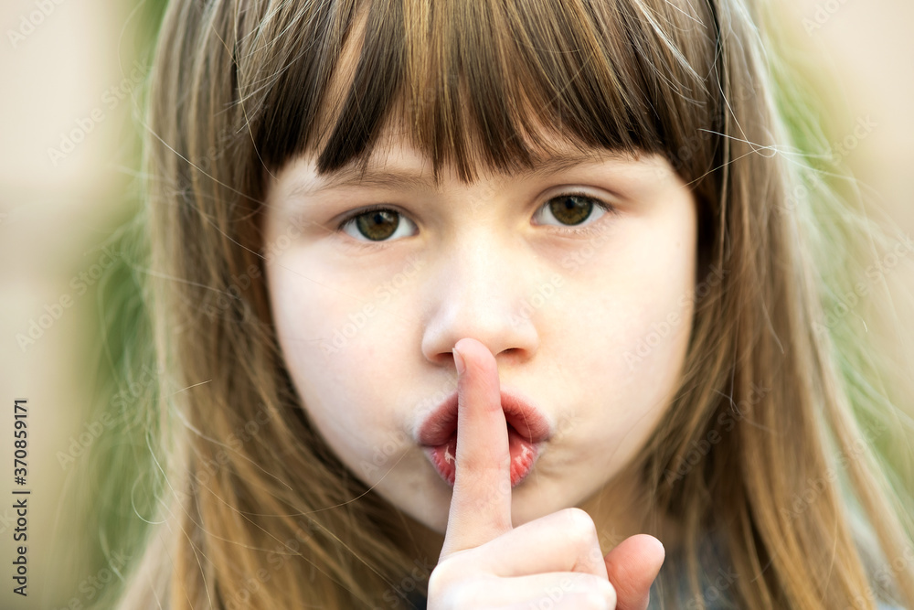 Portrait of pretty child girl with gray eyes and long fair hair holding point finger on her lips with hush sigh. Cute female kid on warm summer day outside. Portrait of pretty child girl with gray eyes and long fair hair holding point finger on her lips with hush sigh. Cute female kid on warm summer day outside.