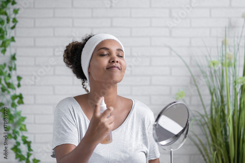 Young woman with eyes closed spraying facial mist at home