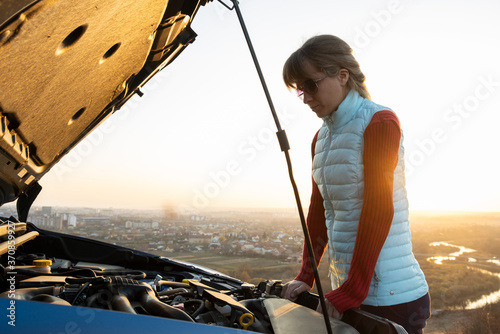 Young woman standing near broken down car with popped up hood having trouble with her vehicle.