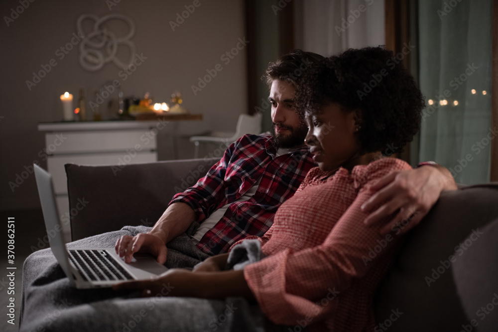 Diverse man and woman hugging and watching film on laptop while resting on couch in evening at home together Diverse man and woman hugging and watching film on laptop while resting on couch in evening at home together