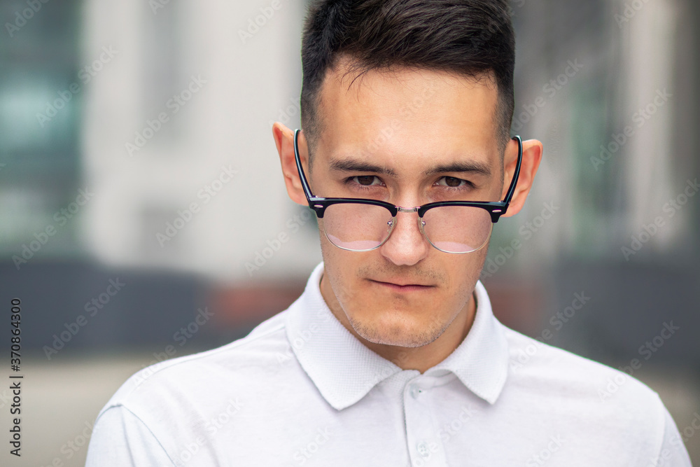 Close up portrait of intelligent smart confident guy, boy in glasses and white polo shirt. Young serious man, student looking at camera over his glasses outdoors. Close up portrait of intelligent smart confident guy, boy in glasses and white polo shirt. Young serious man, student looking at camera over his glasses outdoors.