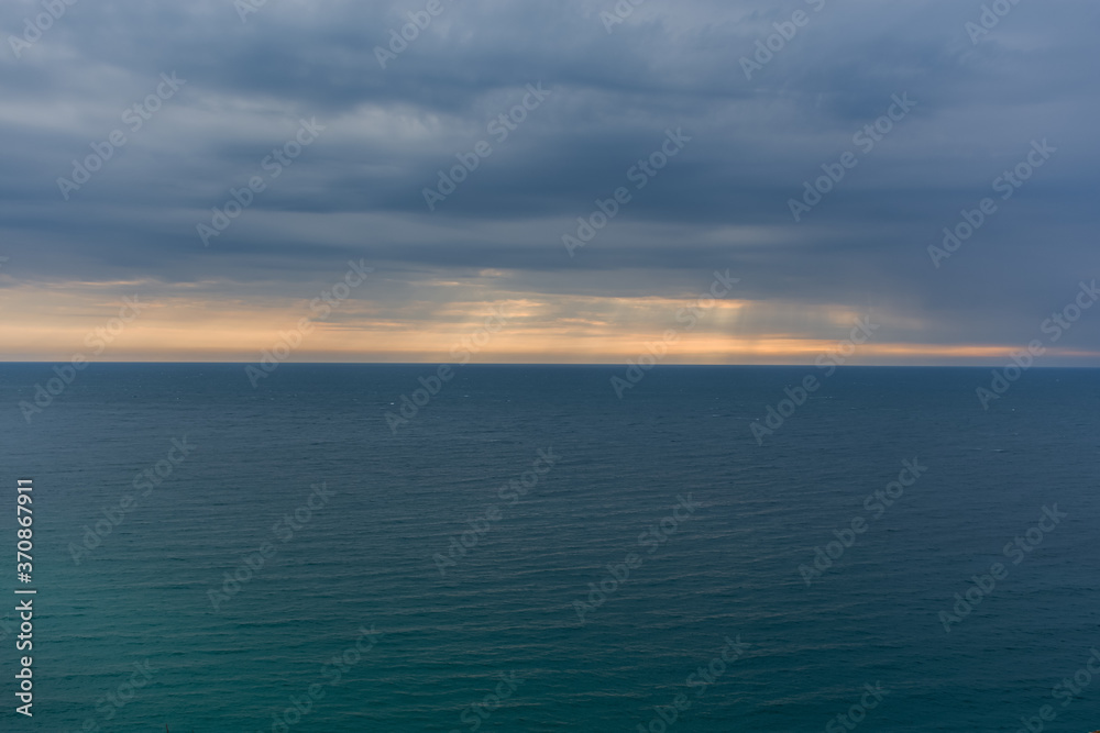 Sea horizon with rays and storm clouds. Calm sea background. Bright blue water and grey rain clouds.