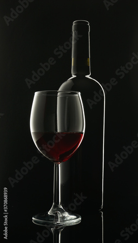 glass goblet with red wine next to a black glass wine bottle on a black background with contour lighting