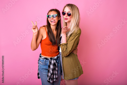 Cheerful positive indoor studio portrait of two funny pretty blonde and brunette woman hugs and looking to each other, surprised emotions, stylish casual outfit, friendship and family concept.
