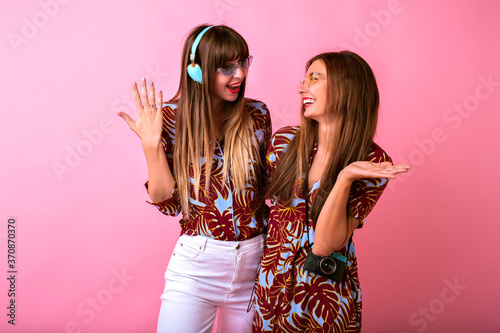 Happy two best friends sister girls having fun showing ok science, color matching tropical print clothes, colorful modern sunglasses, big headphones and vintage camera, students party.