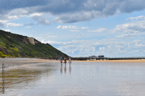 Silhouettes of women and man walking together along north Norfolk coast on low tide summer afternoon at the foot of evergreen hills. UK area of outstanding natural beauty featuring protected landscape