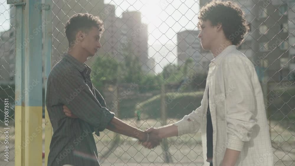 Side view of two young men making peace after conflict outdoors. Portrait of Caucasian and African American friends shaking hands and hugging at sunset and leaving. Multiethnic male friendship.