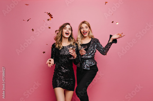 Brown-haired white girl in short dress drinking wine. Indoor photo of spectacular ladies celebrating something with champagne.