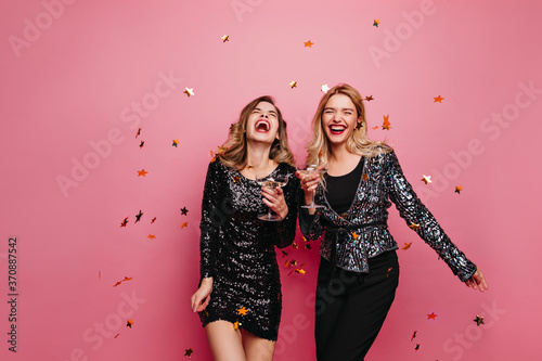Laughing glad ladies celebrating something. Female models in sparkle dresses dancing on pink background.