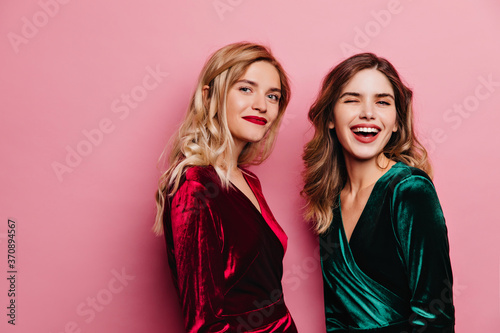 Beautiful elegant women standing on rosy background. Enchanting blonde girl in red velvet dress posing with brunette sister.