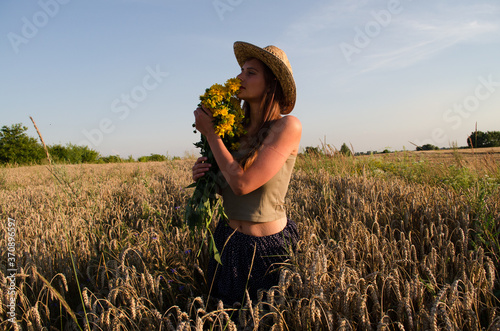 A beautiful woman walks in a field and holds a bouquet of wildflowers,
the girl in the hat smiles and looks into the distance,
a walk among the ears of wheat in August,
natural beauty without makeup