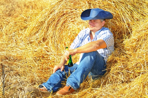 A man cowboy in a hat and jeans and a checkered rough shirt is resting after harvesting  drinking from a bottle of beer on a haystack