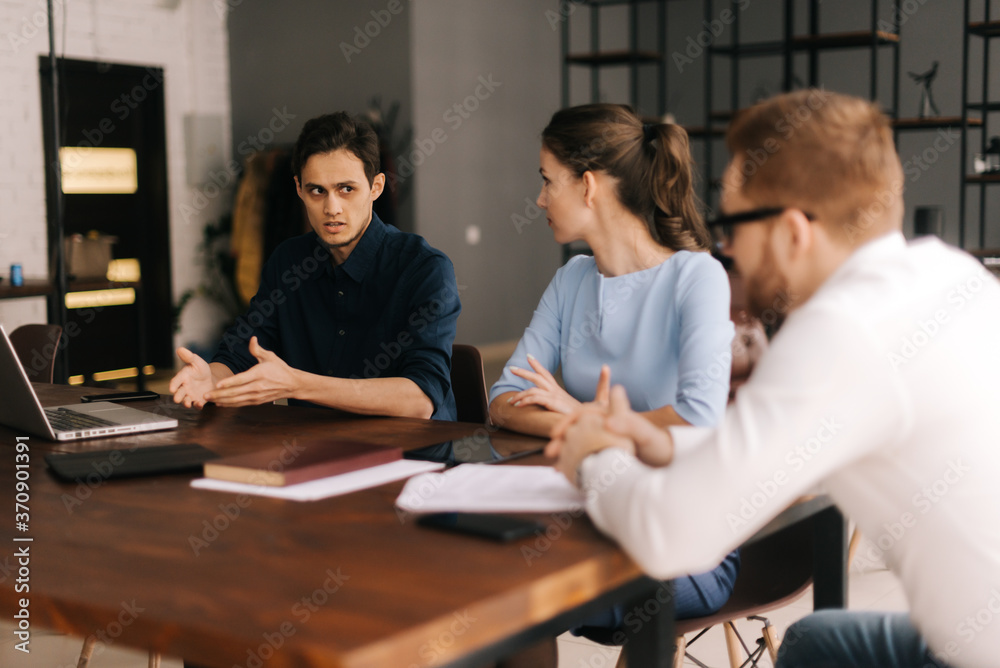 Young business man tells his colleagues about the project in modern office meeting against large panoramic window. Young man and woman listen to the speaker. Concept of office life. Young business man tells his colleagues about the project in modern office meeting against large panoramic window. Young man and woman listen to the speaker. Concept of office life.