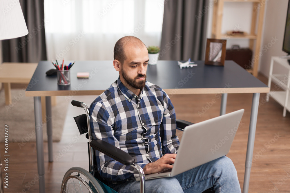 Businessman in wheelchair working remotely from home on laptop. Hopeful, handicapped entrepreneur in wheelchair communicating online with business partners sitting in his apartment.