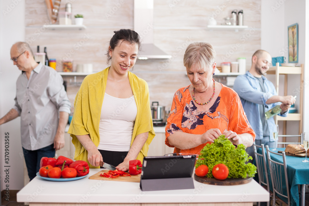 Mother and daughter following online recipe on tablet for healthy salad. Husband smiling while holding bottle of wine. Senior man opening refrigerator door in kitchen. Mother and daughter following online recipe on tablet for healthy salad. Husband smiling while holding bottle of wine. Senior man opening refrigerator door in kitchen.