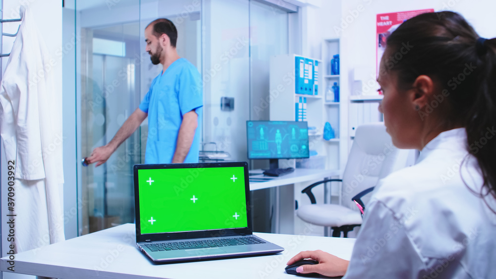 Medical worker in white coat using laptop with green screen in hospital cabinet. Nurse wearing blue uniform opening glass door. Medic wearing uniform using notebook with chroma key on display in