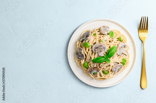 Spaghetti with creamy sauce, champignons and green basil on a light blue background. Vegetarian Italian pasta with mushrooms in a white plate and a golden fork. Top view, copy space.