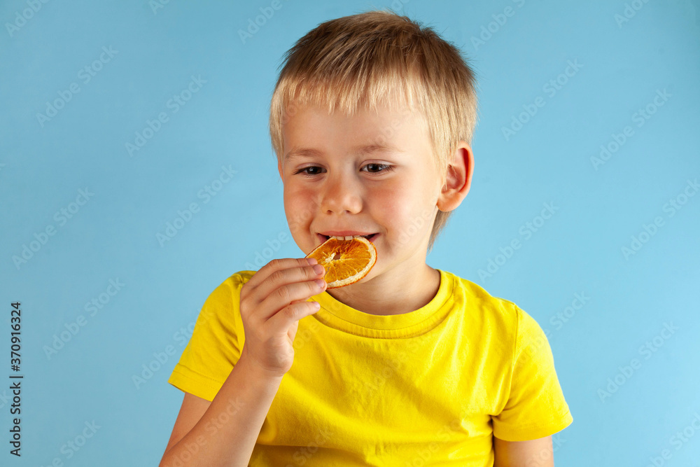 A blond boy is smiling and eating dried orange chips on a blue background. The concept of healthy food for children, a healthy snack. A blond boy is smiling and eating dried orange chips on a blue background. The concept of healthy food for children, a healthy snack.