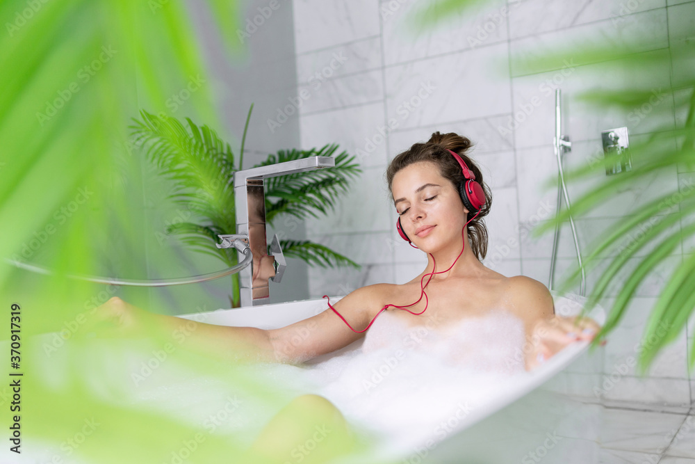 © deniskomarov - Beautiful relaxed girl wearing headphones enjoy listening to music with eyes closed while taking bath with soap foam, warm water. Stress relief, mental rest. Female portrait with view through leaves © deniskomarov - Beautiful relaxed girl wearing headphones enjoy listening to music with eyes closed while taking bath with soap foam, warm water. Stress relief, mental rest. Female portrait with view through leaves