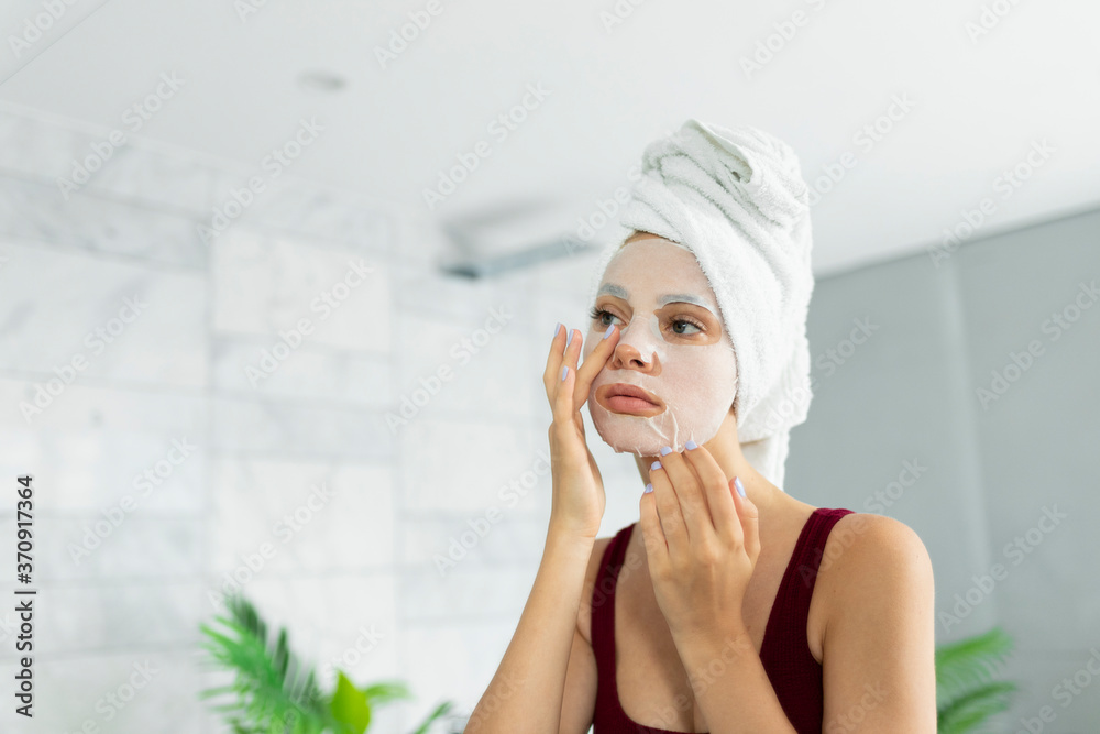 Beautiful young woman applying in sheet face mask wearing bath towel on head looking at mirror reflection. Cosmetological procedure for skin cleansing and moisturizing. Professional skincare Beautiful young woman applying in sheet face mask wearing bath towel on head looking at mirror reflection. Cosmetological procedure for skin cleansing and moisturizing. Professional skincare