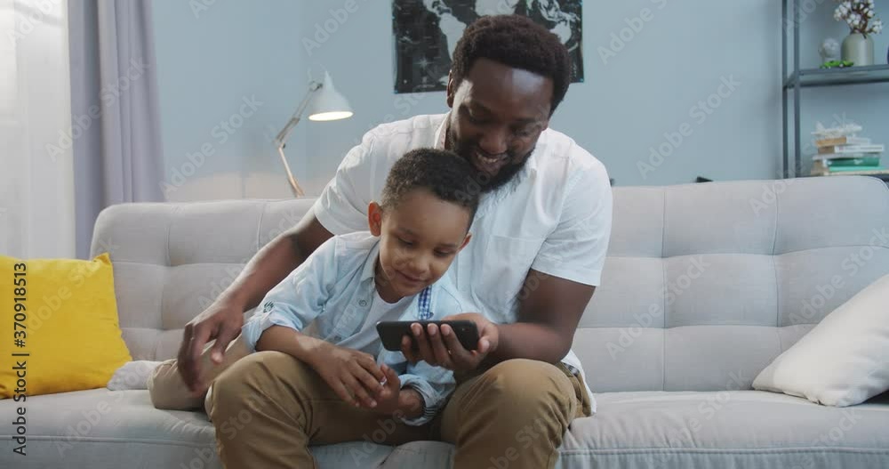 Happy African American male watching video on smartphone with small cute son and imitating moves while sitting on sofa at home. Handsome joyful father showing something on cellphone to little kid Happy African American male watching video on smartphone with small cute son and imitating moves while sitting on sofa at home. Handsome joyful father showing something on cellphone to little kid