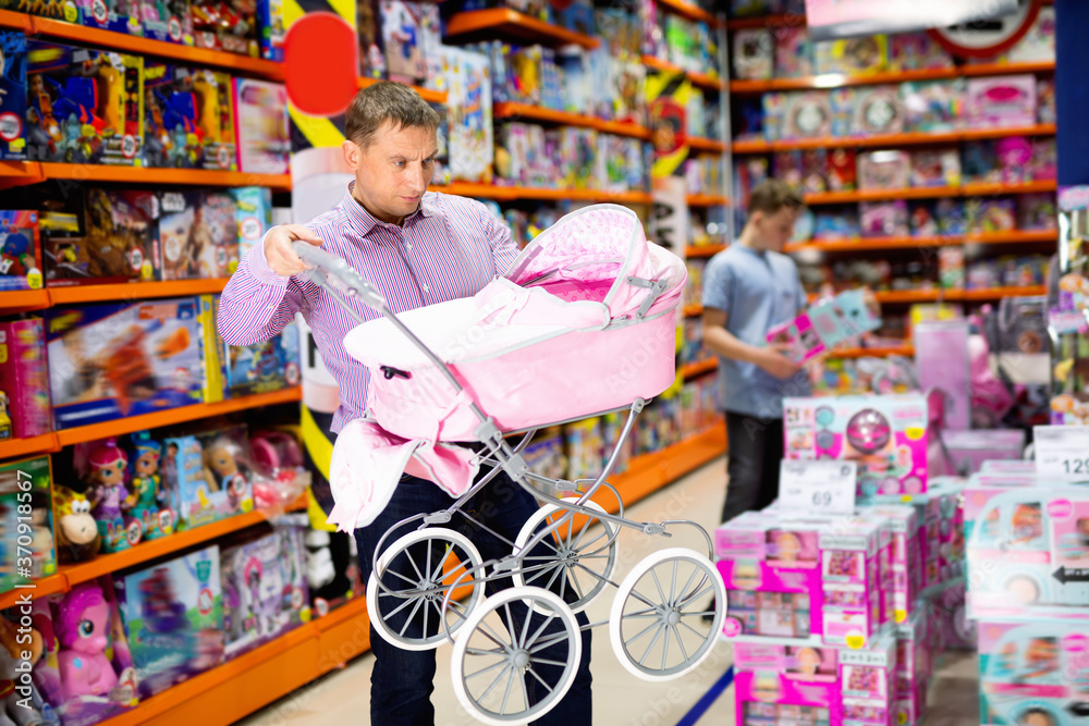 Caring glad cheerful positive smiling father choosing pink baby doll stroller for gift to his daughter in children toy store