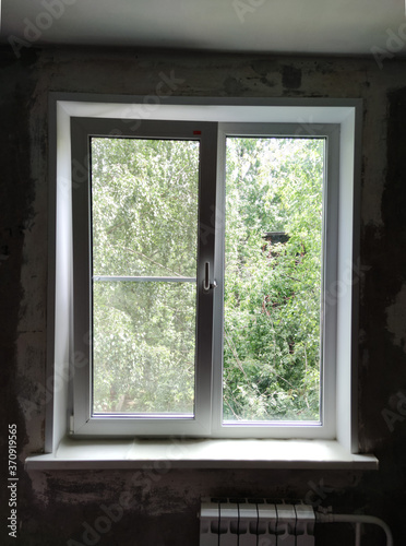 Plastic white window with a view of green trees in summer.