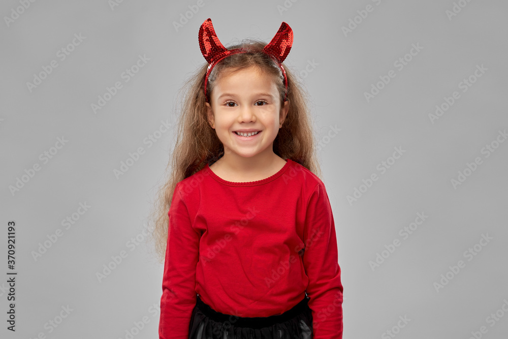 halloween, holiday and childhood concept - smiling girl in party costume and red devil's horns over grey background