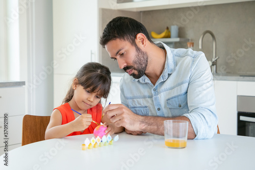 Handsome dad painting Easter egg with daughter at kitchen table. Cute girl in red shirt holding brush  sitting and drawing with young Latin father. Family time  childhood and holiday concept