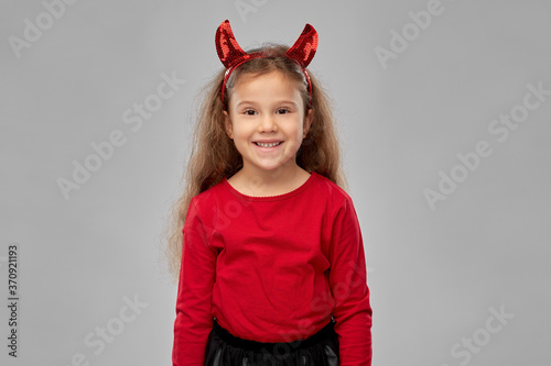 halloween, holiday and childhood concept - smiling girl in party costume and red devil's horns over grey background