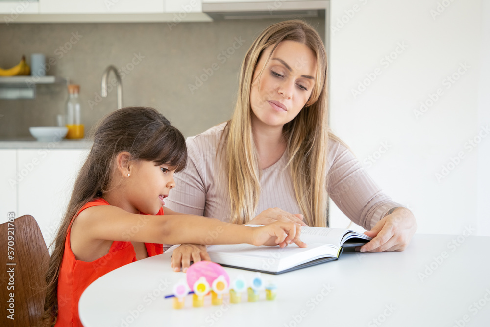 Cute little girl pointing at text and learning with mom. Attractive young blonde mother teaching little daughter and reading book with her in kitchen. Family time, childhood and education concept Cute little girl pointing at text and learning with mom. Attractive young blonde mother teaching little daughter and reading book with her in kitchen. Family time, childhood and education concept