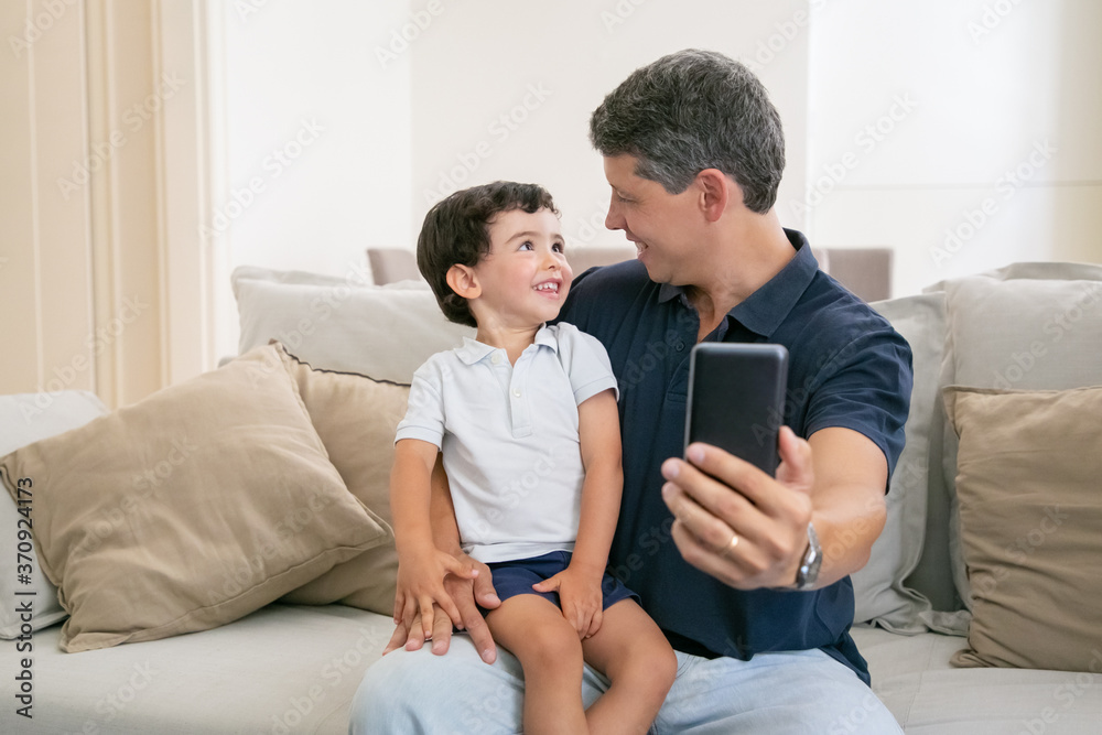 Happy dad and little son enjoying time together, sitting on couch at home, chatting, laughing and taking selfie. Medium shot, front view. Family or technology concept Happy dad and little son enjoying time together, sitting on couch at home, chatting, laughing and taking selfie. Medium shot, front view. Family or technology concept