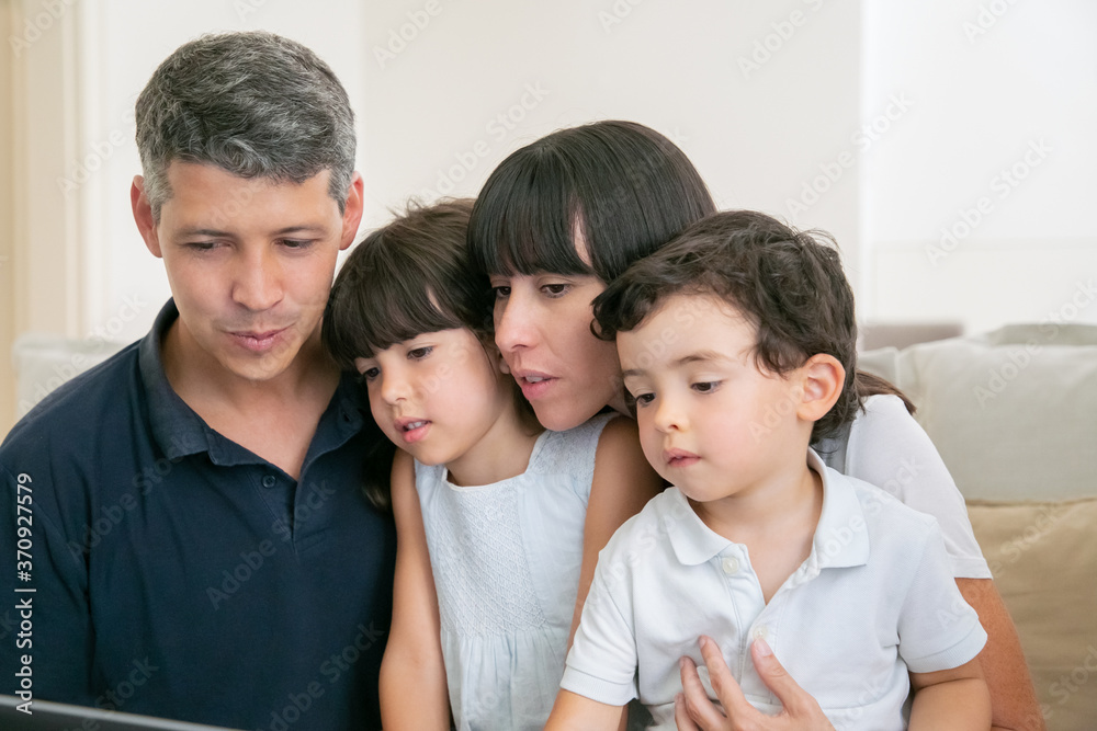 Parent couple and two kids using computer for video call, sitting on couch together, looking at display. Medium shot. Communication or entertainment internet concept Parent couple and two kids using computer for video call, sitting on couch together, looking at display. Medium shot. Communication or entertainment internet concept