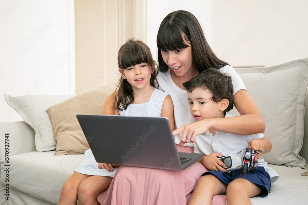 Positive excited mother embracing two kids and pointing at laptop display. Family sitting on couch at home and watching movie. Medium shot. Communication or entertainment concept Positive excited mother embracing two kids and pointing at laptop display. Family sitting on couch at home and watching movie. Medium shot. Communication or entertainment concept