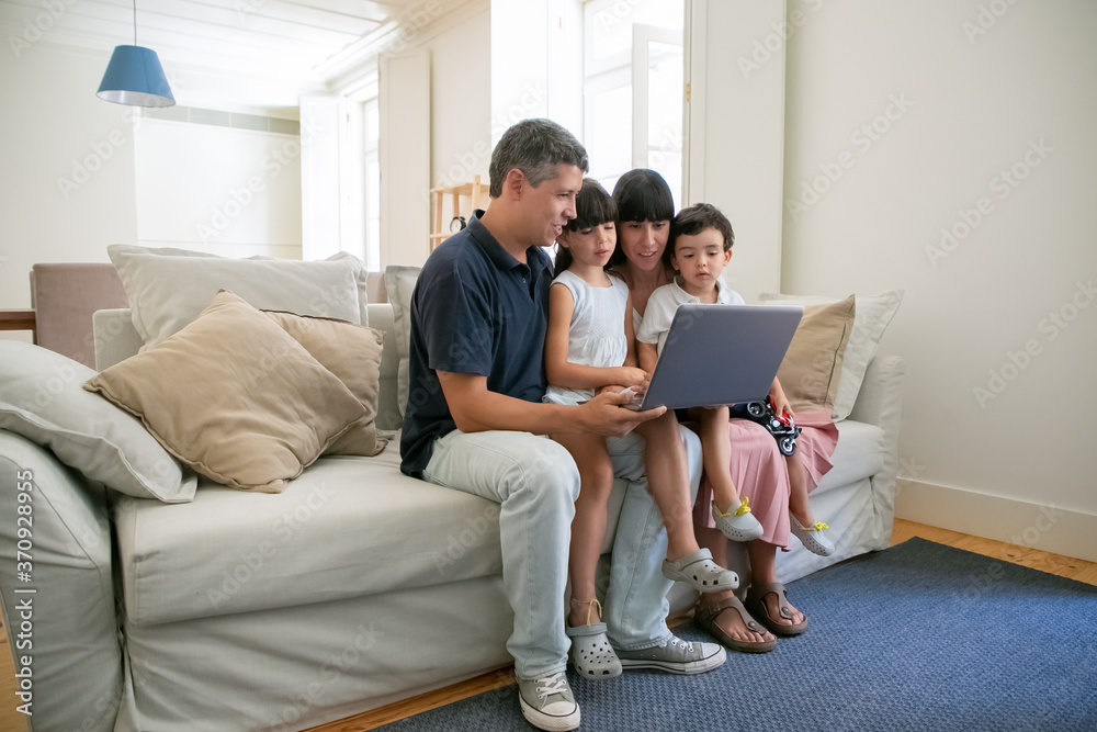 Family couple holding kids on lap, sitting on couch all together, watching movie or video on laptop at home. Full length. Communication or entertainment concept