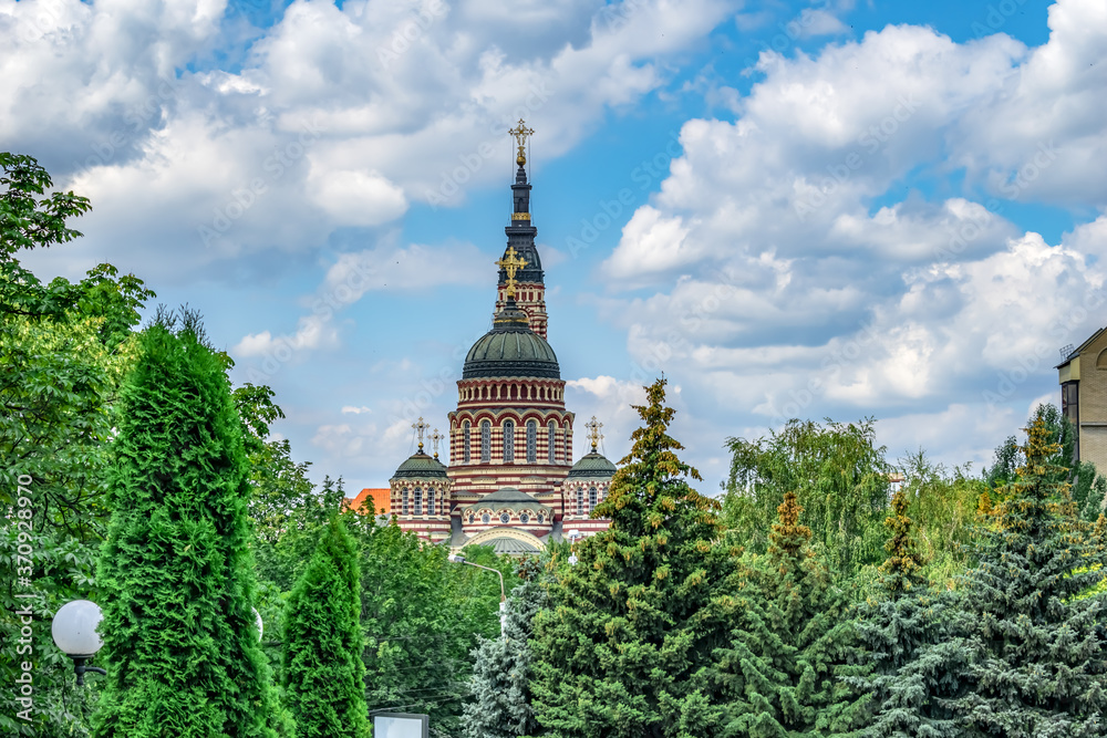 Domes with crosses of Annunciation Cathedral in Kharkiv (Ukraine) against a blue cloudy sky. Park with green summer trees against the background of a red-beige striped Orthodox church on a sunny day
