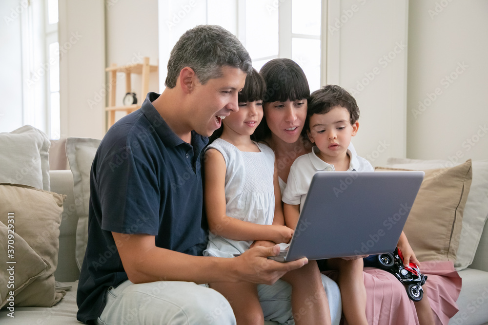 Excited cheerful parents couple holding kids on lap, sitting on couch all together, watching movie or video on laptop at home, staring at display. Medium shot. Communication or entertainment concept Excited cheerful parents couple holding kids on lap, sitting on couch all together, watching movie or video on laptop at home, staring at display. Medium shot. Communication or entertainment concept