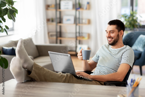 technology, remote job and lifestyle concept - happy smiling man with laptop computer drinking coffee and resting feet on table at home office