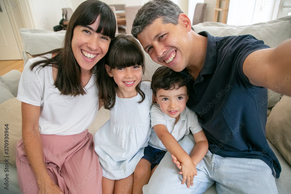 Joyful parents and two kids sitting on couch at home together, taking selfie, dad holding camera in hand. Front view, pov shot. Family photo concept Joyful parents and two kids sitting on couch at home together, taking selfie, dad holding camera in hand. Front view, pov shot. Family photo concept