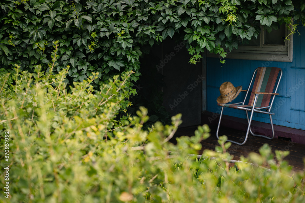 A summer hat and a folding chair surrounded by green vine leaves on a wooden deck. Cottage aesthetics. Vacation outside the city. Warm summer day. A summer hat and a folding chair surrounded by green vine leaves on a wooden deck. Cottage aesthetics. Vacation outside the city. Warm summer day.